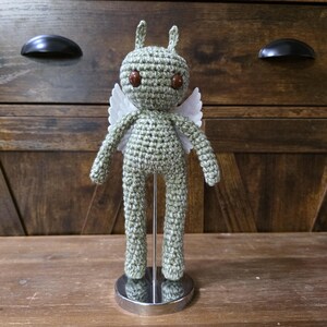 May include: A handmade crochet doll in a light green color, featuring brown eyes, small white wings, and a pair of small antennae. The doll is standing on a silver stand. The background is a wooden cabinet.