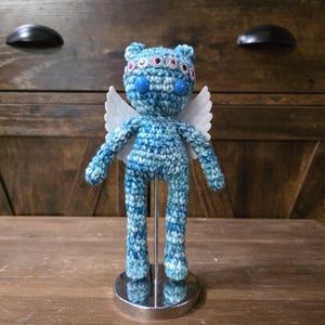 May include: A handmade blue and white crochet doll with a bear-like face, large blue eyes, and a jeweled headband. The doll has white wings and is mounted on a silver stand. The background features a wooden cabinet.