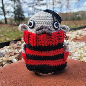 May include: A gray crocheted plush toy with black hair, large eyes, and a red and black striped sweater.