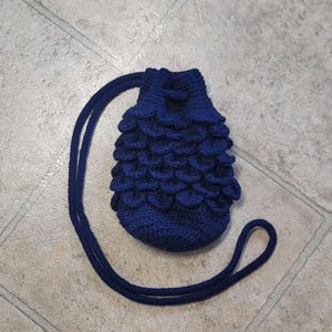 May include: A small, dark blue crocheted drawstring bag with a long strap. The bag is made with a textured, scale-like pattern.