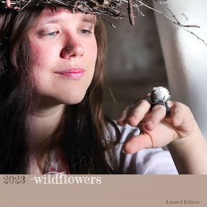 May include: A close-up of a ring with a white, textured design and a dark band. The ring is on a person's finger. The image also includes the text "2023 - wildflowers" and "Limited Edition".