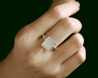 May include: A silver ring with a square-shaped design featuring a geometric pattern. The ring is worn on the index finger of a hand.