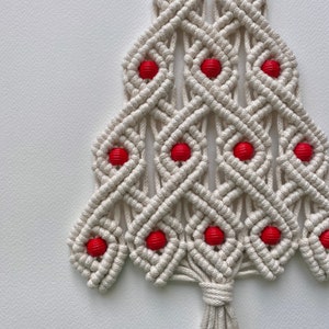 May include: A white macrame Christmas tree wall hanging with red beads. The tree is made with a diamond pattern and has a tassel at the bottom.