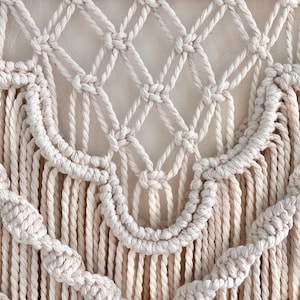 K&ouml;nnte beinhalten: Ein wei&szlig;er Makramee-Wandbehang mit geometrischem Muster und Fransen. Der Wandbehang ist aus Baumwollschnur gefertigt und hat einen Boho-Stil.