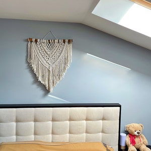 May include: A white macrame wall hanging with a wooden dowel rod at the top, hanging above a tufted headboard with a brown and white color scheme. A brown teddy bear with a red bow is sitting on the floor next to the bed.