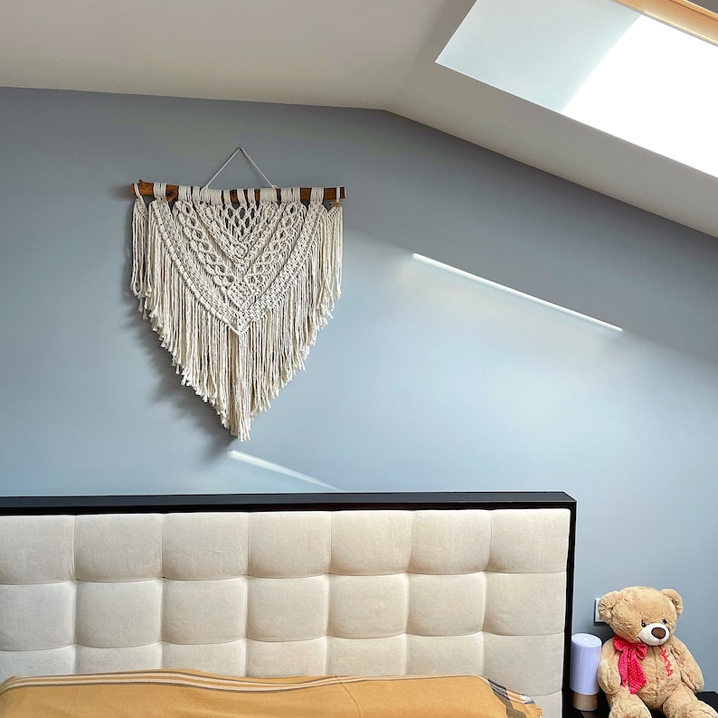 May include: A white macrame wall hanging with a wooden dowel rod at the top, hanging above a tufted headboard with a brown and white color scheme. A brown teddy bear with a red bow is sitting on the floor next to the bed.