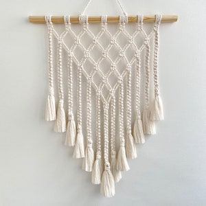 May include: A white macrame wall hanging with a diamond pattern and fringe tassels. The hanging is made of cotton cord and is attached to a wooden dowel.