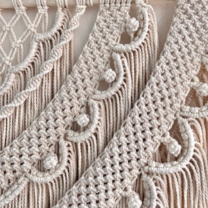 K&ouml;nnte beinhalten: Ein beigefarbener Makramee-Wandbehang mit geometrischem Muster und Fransen. Der Wandbehang ist aus nat&uuml;rlichem Baumwollgarn gefertigt und hat einen Boho-Stil.