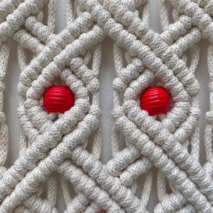 May include: Close-up of a macrame wall hanging with a geometric pattern. The hanging is made with white cord and features two red wooden beads.