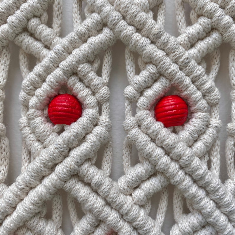 May include: Close-up of a macrame wall hanging with a geometric pattern. The hanging is made with white cord and features two red wooden beads.