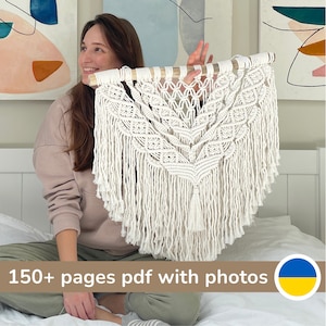 May include: A woman is holding up a large white macrame wall hanging with a wooden dowel at the top. The wall hanging has a fringe and a tassel at the bottom. The text "150+ pages pdf with photos" is at the bottom of the image. A Ukrainian flag is in the bottom right corner.