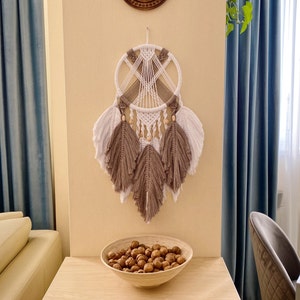 May include: A white and brown macrame dream catcher with wooden beads hangs on a wall with a light blue curtain. The dream catcher has a circular frame with a web-like design and feathers.