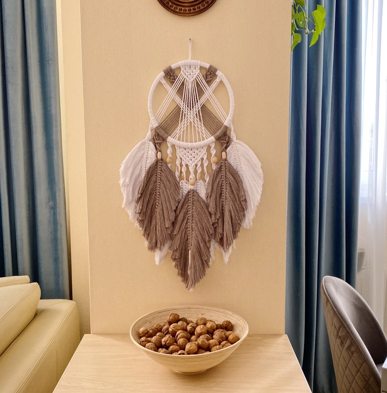 May include: A white and brown macrame dream catcher with wooden beads hangs on a wall with a light blue curtain. The dream catcher has a circular frame with a web-like design and feathers.