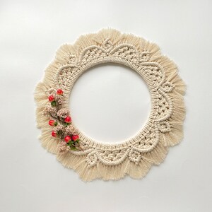 May include: A cream-colored macrame wreath with a fringe border and a cluster of red berries.