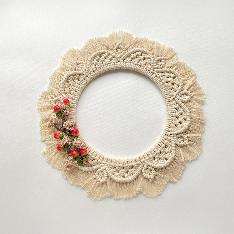 May include: A cream-colored macrame wreath with a fringe border and a cluster of red berries.