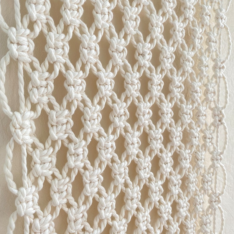 Może przedstawiać: Biała wisząca ozdoba ścienna wykonana techniką makramy z rombowym wzorem. Makrama wykonana jest z grubego sznurka bawełnianego i ma naturalny, boheński styl.