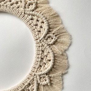 May include: A cream-colored macrame wall hanging with a circular shape and a fringe border. The macrame is made with a thick, natural fiber rope.
