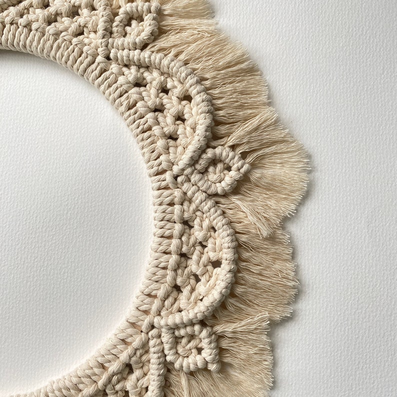 May include: A cream-colored macrame wall hanging with a circular shape and a fringe border. The macrame is made with a thick, natural fiber rope.
