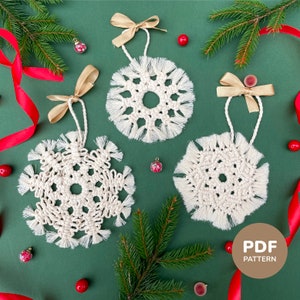 Könnte beinhalten: Drei handgefertigte, cremefarbene Makramee-Schneeflocken-Ornamente mit Fransen, gebunden mit goldenem Band und an weißen Kordeln hängend. Die Ornamente befinden sich auf einem grünen Hintergrund mit roten Beeren, Tannennadeln und einem roten Band. Ein PDF-Muster-Symbol befindet sich unten rechts.