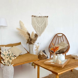May include: A bohemian living room with a macrame wall hanging, a wicker chair, a wooden coffee table, and a couch with throw pillows. The room is decorated with dried pampas grass and other natural elements.