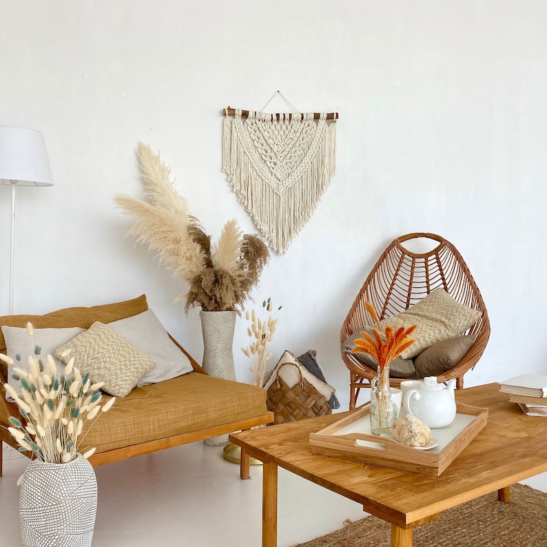 May include: A bohemian living room with a macrame wall hanging, a wicker chair, a wooden coffee table, and a couch with throw pillows. The room is decorated with dried pampas grass and other natural elements.