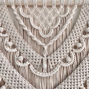K&ouml;nnte beinhalten: Ein beigefarbener Makramee-Wandbehang mit geometrischem Muster und Fransen. Der Wandbehang ist aus Naturfasern gefertigt und hat einen Boho-Stil.