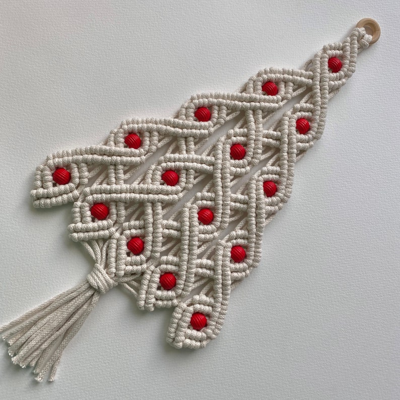 May include: A white macrame Christmas tree wall hanging with red beads. The tree has a tassel at the bottom.