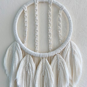 May include: A white macrame dream catcher with a circular hoop and multiple white feather-like tassels hanging from the bottom.