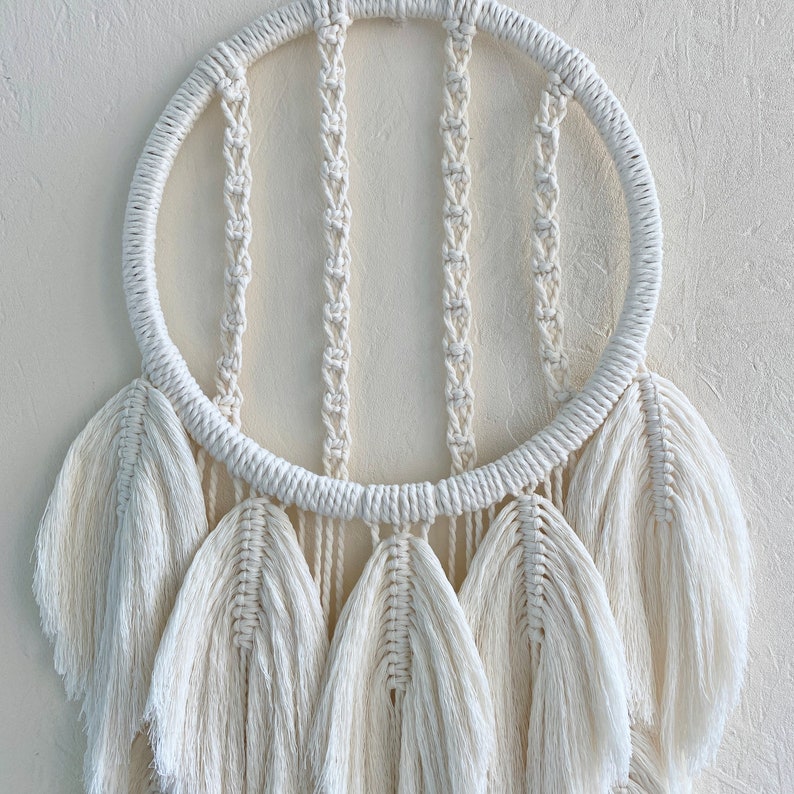 May include: A white macrame dream catcher with a circular hoop and multiple white feather-like tassels hanging from the bottom.