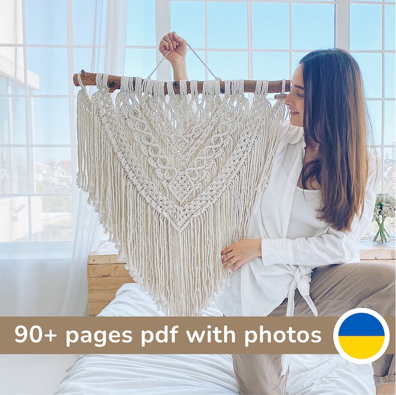 May include: A woman holds up a large, white macrame wall hanging with a geometric pattern. The hanging is made of cotton cord and has a wooden dowel at the top. The woman is wearing a white shirt and beige pants. The text "90+ pages pdf with photos" is visible at the bottom of the image.