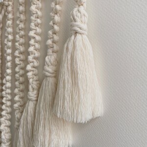 May include: Close-up of a white macrame wall hanging with multiple tassels. The tassels are made of thick, soft, white yarn.