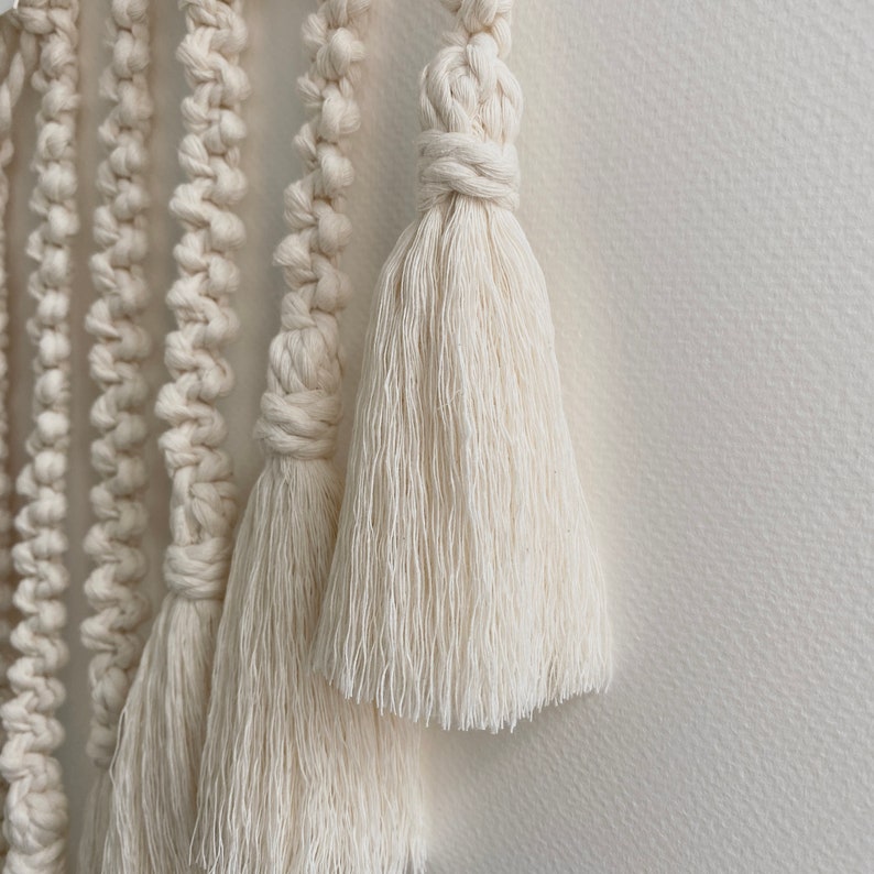 May include: Close-up of a white macrame wall hanging with multiple tassels. The tassels are made of thick, soft, white yarn.