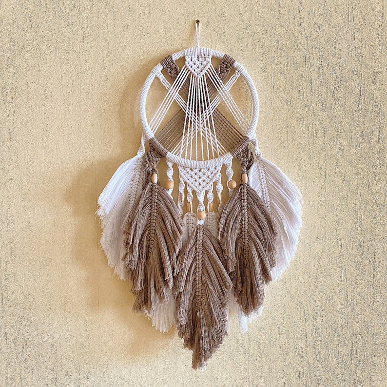 May include: A white and brown dream catcher with wooden beads and feathers. The dream catcher is hanging from a white wall.
