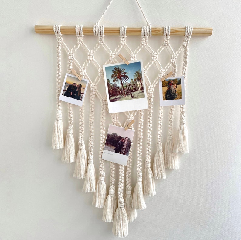 May include: A white macrame wall hanging with a wooden dowel rod at the top. The macrame has a diamond pattern and features four photo spaces with photos of people and a cityscape. The macrame has tassels hanging from the bottom.