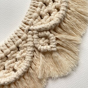 May include: A close-up of a cream-colored macrame wall hanging with a fringe border. The macrame is made with a knotting technique that creates a geometric pattern.