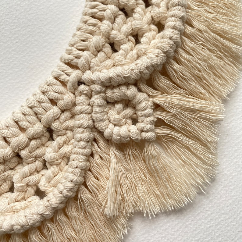 May include: A close-up of a cream-colored macrame wall hanging with a fringe border. The macrame is made with a knotting technique that creates a geometric pattern.