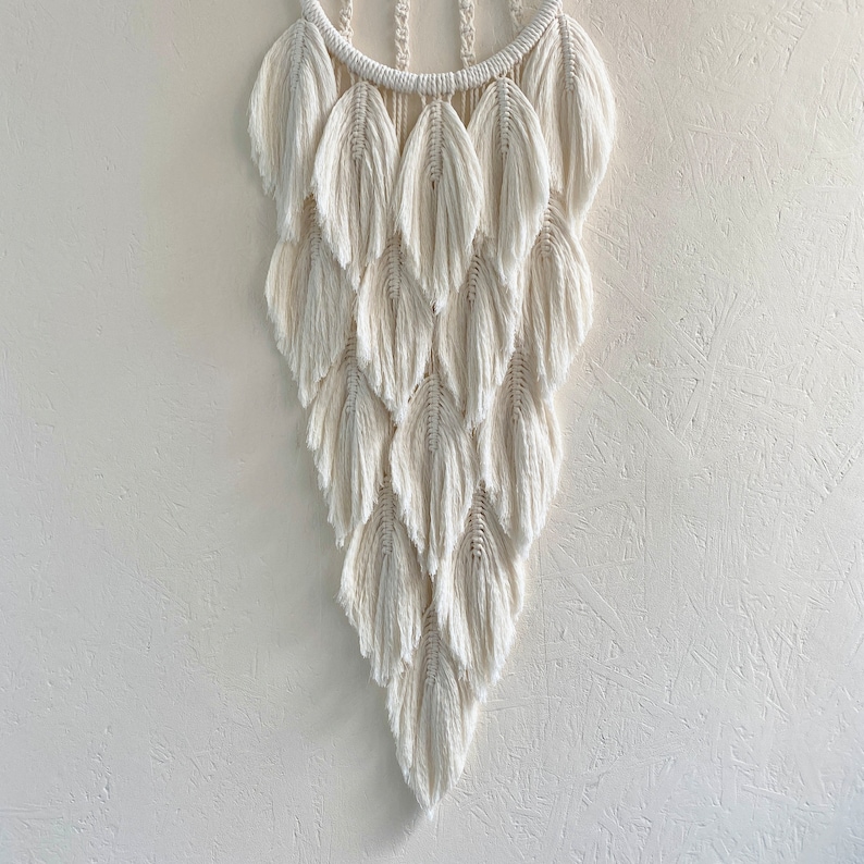 May include: A white macrame wall hanging with a circular top and a cascading design of feather-shaped knots.