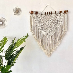 May include: A white macrame wall hanging with a geometric pattern and fringe. The hanging is made with natural cotton cord and is attached to a wooden branch.