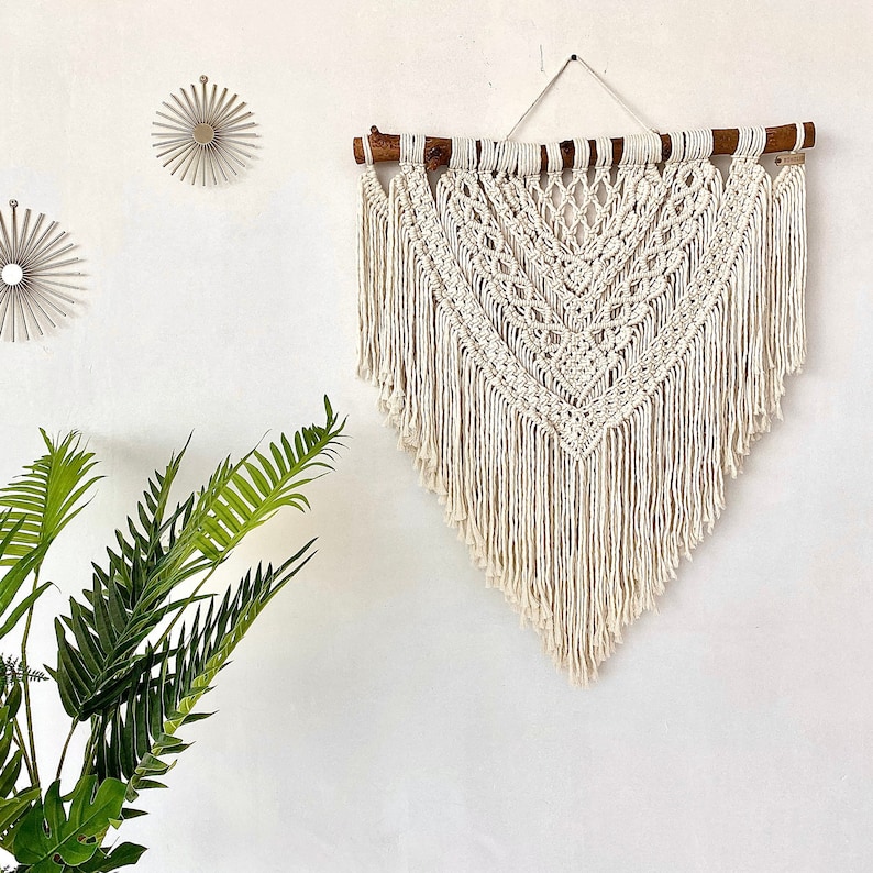 May include: A white macrame wall hanging with a geometric pattern and fringe. The hanging is made with natural cotton cord and is attached to a wooden branch.