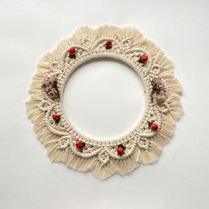 May include: A cream-colored macrame wreath with a circular shape and a fringe border. The wreath is decorated with small red berries and dried flowers.