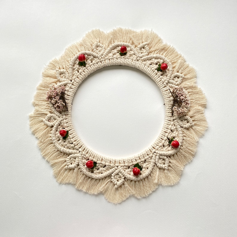 May include: A cream-colored macrame wreath with a circular shape and a fringe border. The wreath is decorated with small red berries and dried flowers.