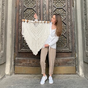 May include: A white macrame wall hanging with a wooden dowel at the top. The hanging is in the shape of a heart with fringe at the bottom. A person is holding the hanging in front of a wooden door.