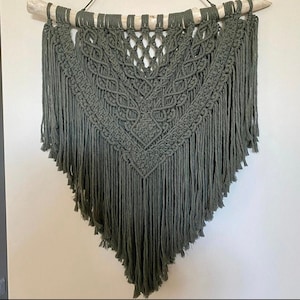 May include: A large, green macrame wall hanging with a geometric pattern and fringe. The hanging is made with natural fibers and is attached to a wooden branch.