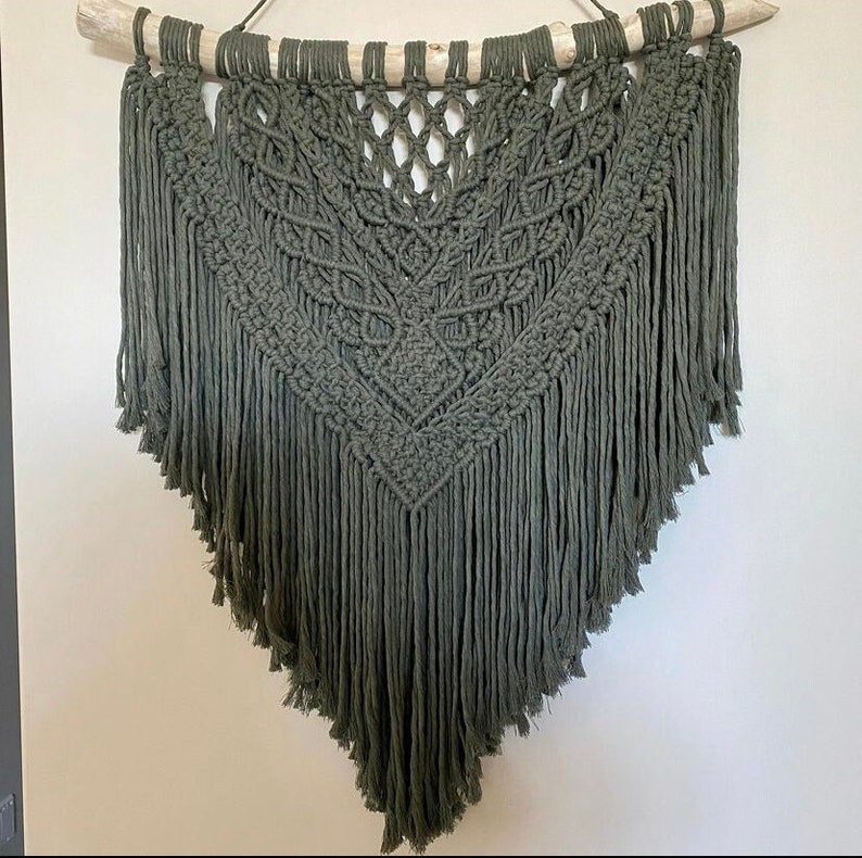 May include: A large, green macrame wall hanging with a geometric pattern and fringe. The hanging is made with natural fibers and is attached to a wooden branch.
