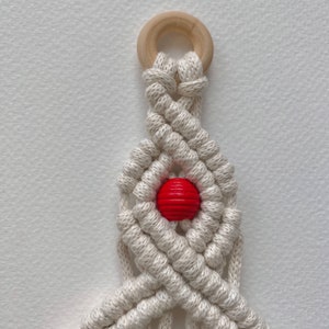 May include: A white macrame plant hanger with a wooden ring at the top and a red wooden bead in the center.