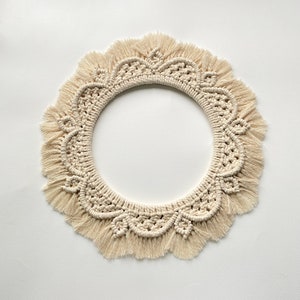 May include: A cream-colored macrame wall hanging with a circular shape and a fringe border. The macrame is made with a knotting technique and has a delicate, intricate design.
