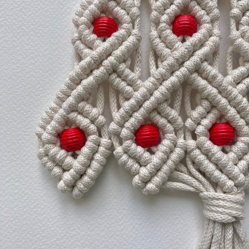 May include: A white macrame wall hanging in the shape of a Christmas tree with red wooden beads.