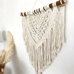 May include: A white macrame wall hanging with a wooden dowel at the top. The hanging features a geometric pattern with fringe at the bottom. The dowel has a small tag with the word "BOHO" written on it.