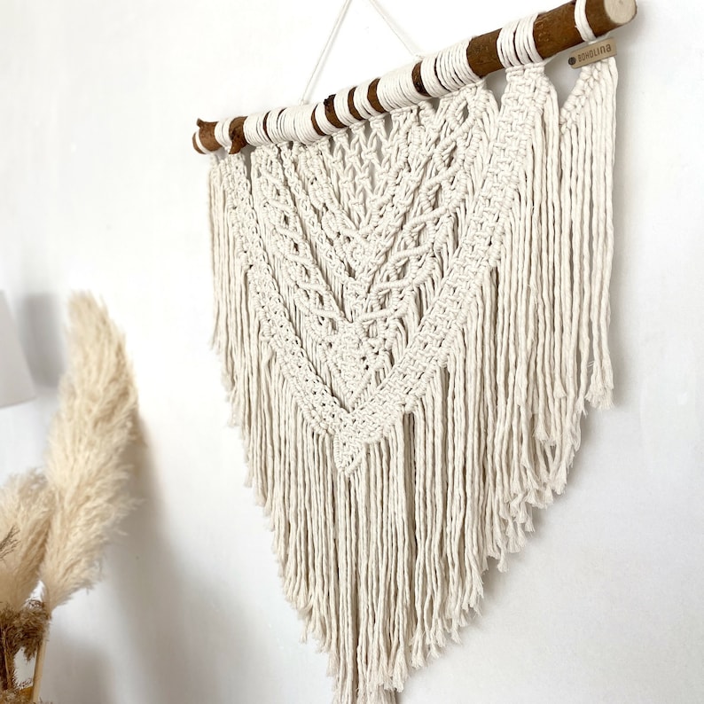 May include: A white macrame wall hanging with a wooden dowel at the top. The hanging features a geometric pattern with fringe at the bottom. The dowel has a small tag with the word "BOHO" written on it.