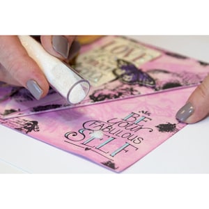 Op de afbeelding: Een doorzichtige tube strooit witte glitter op een roze kaart. De kaart bevat de woorden "BE YOUR FABULOUS SELF" in turquoise en zwart, samen met vlinder- en bloemmotieven. Het woord "LOVE" is ook zichtbaar.